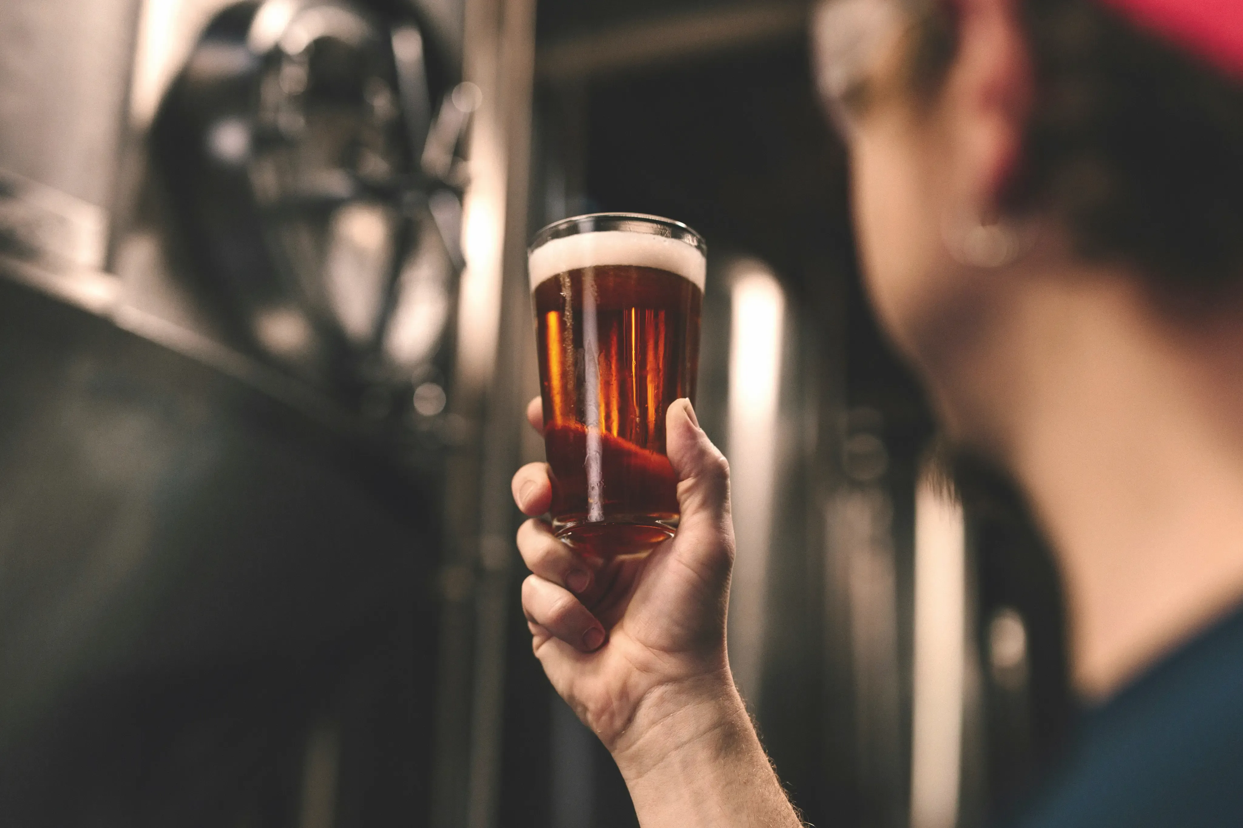 A person holds up a glass of amber ale in a brewery, with blurred stainless steel fermentation tanks in the background.